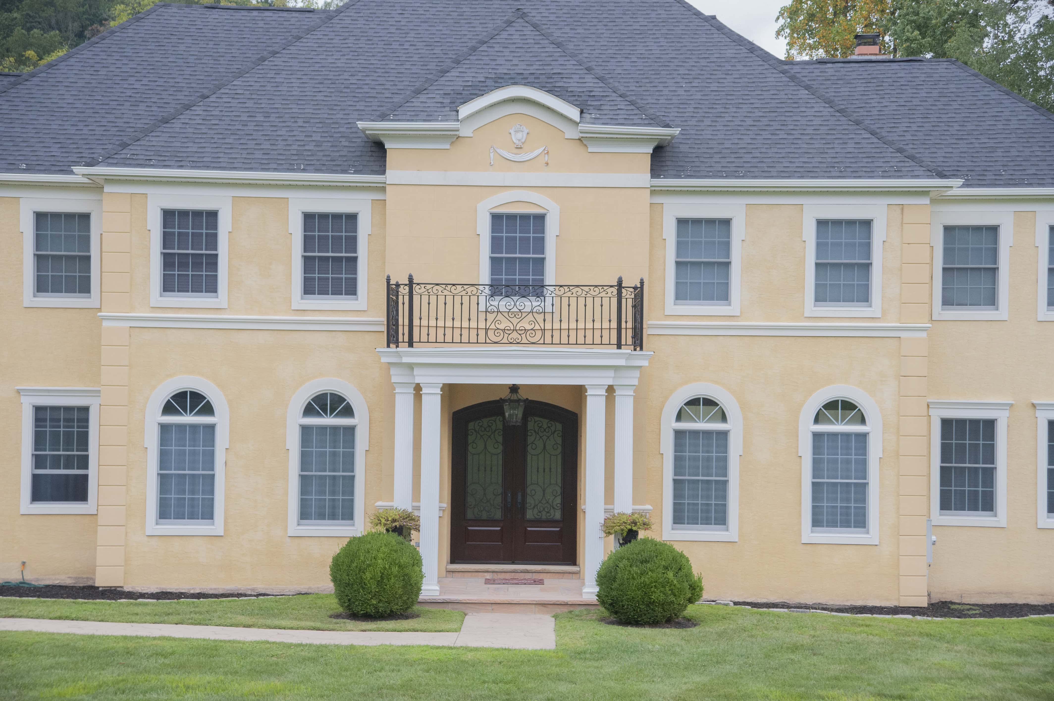 A large two-story yellow house with white trim, multiple windows, a dark gray roof, a small balcony above the main entrance, and neatly trimmed bushes lining the front walkway.