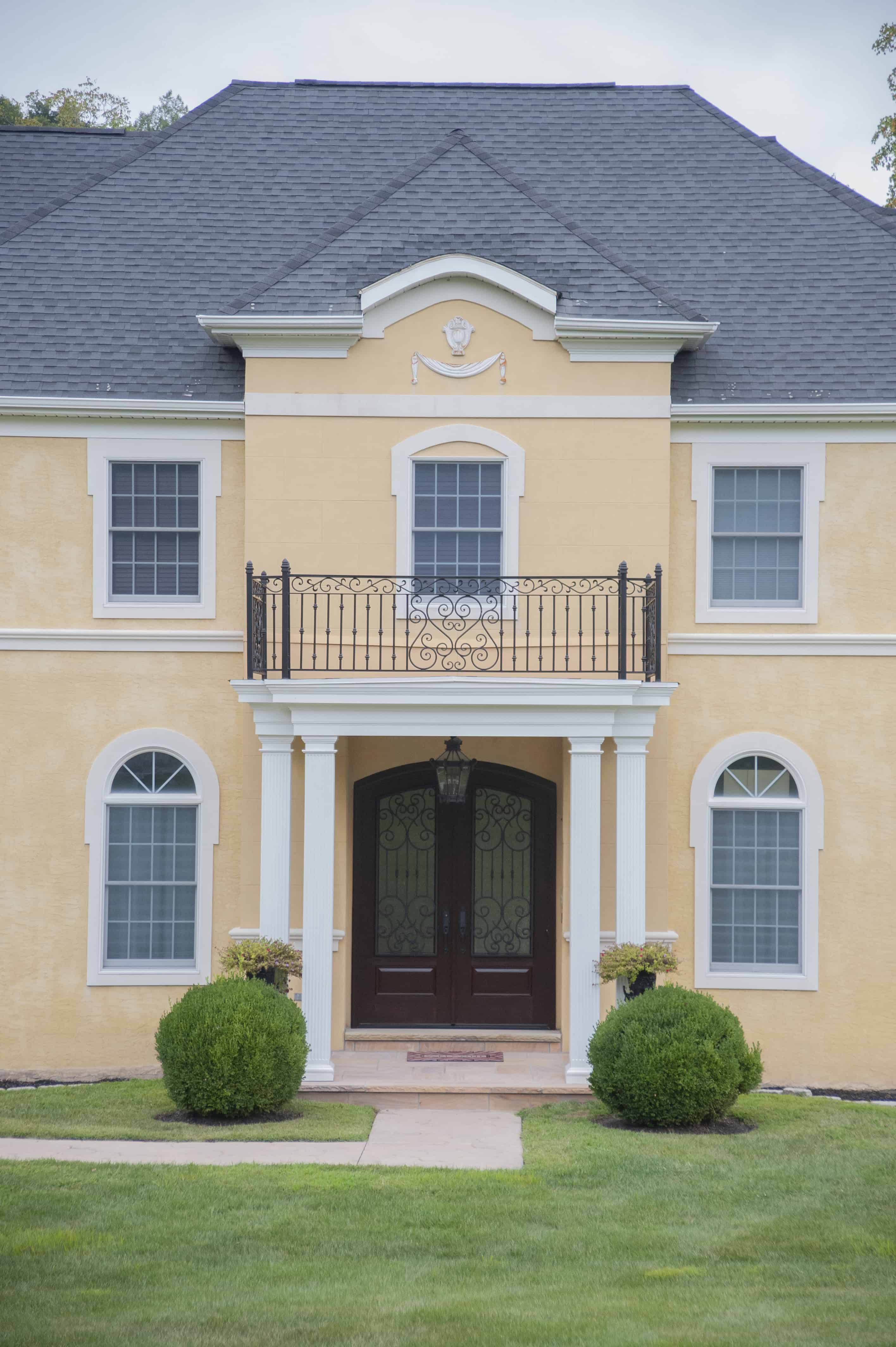 A two-story yellow house with a dark gray roof, white columns, arched windows, and a decorative black balcony above double front doors, set in a green lawn with neatly trimmed bushes.