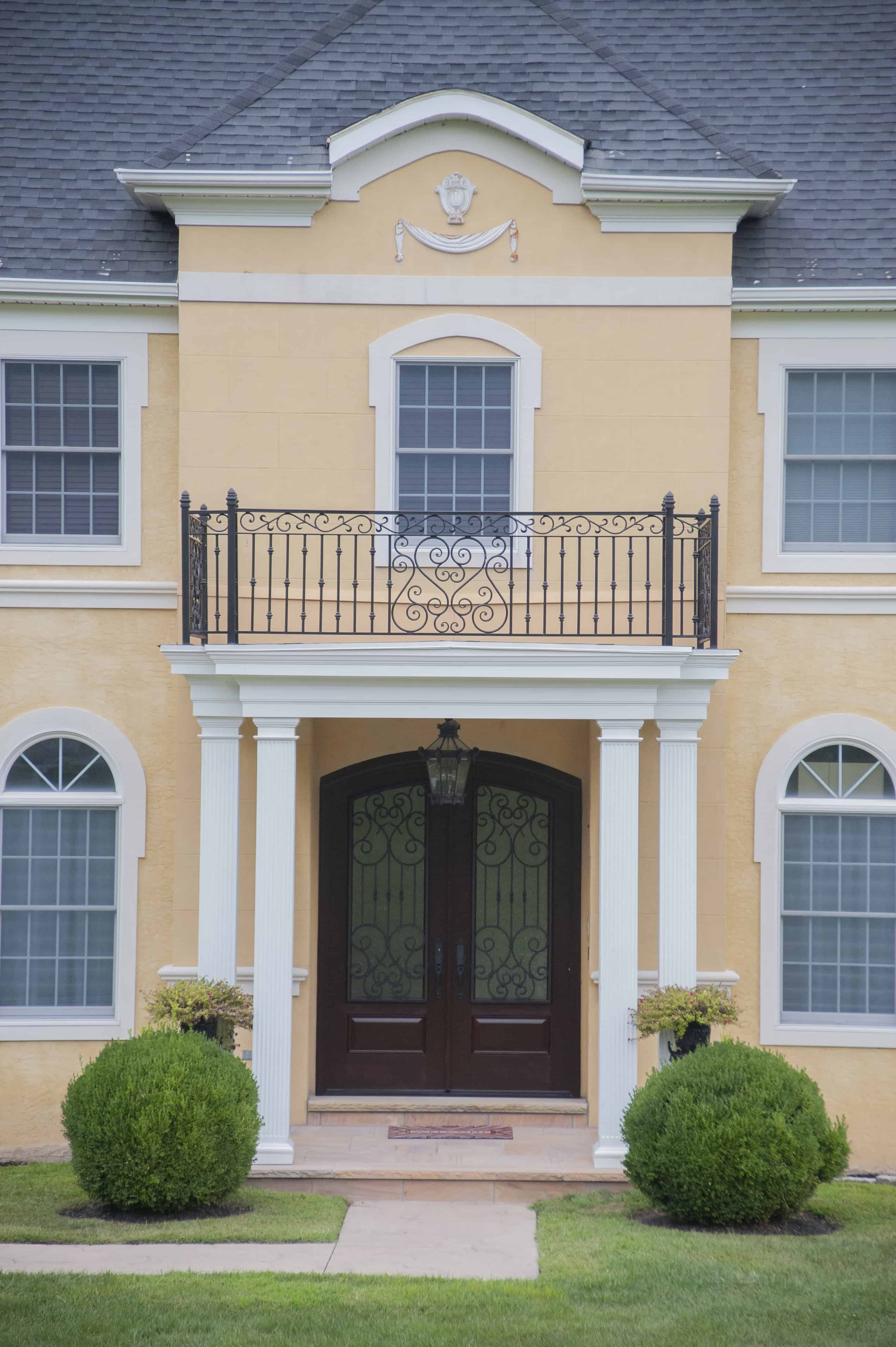 A two-story yellow house with white trim, arched windows, a black wrought-iron balcony, and a double front door with white columns and manicured bushes on either side of the entrance.