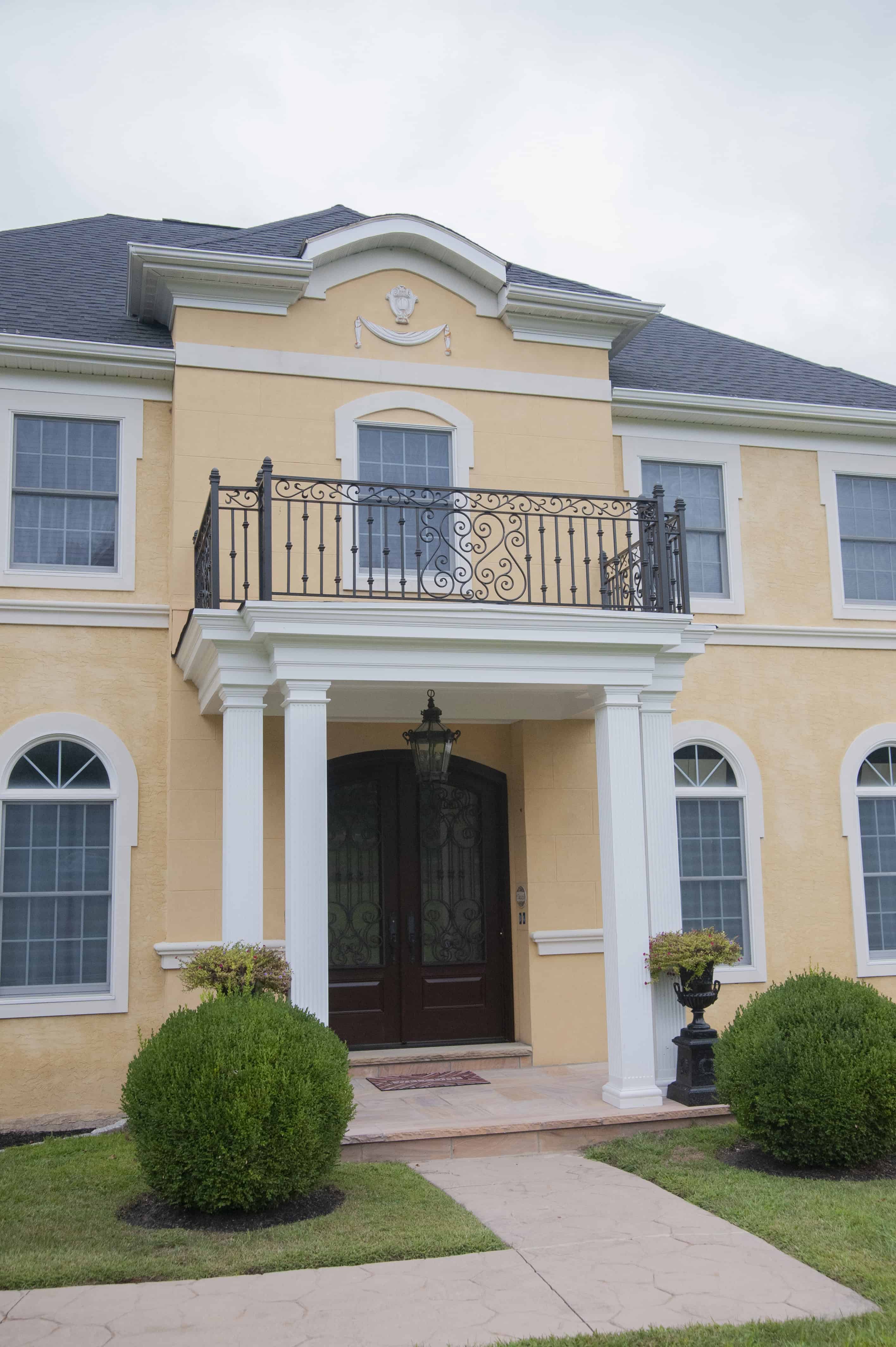 A two-story yellow house with white trim, arched windows, a black wrought-iron balcony, double front doors, and decorative columns at the entrance, surrounded by green shrubs and a small lawn.