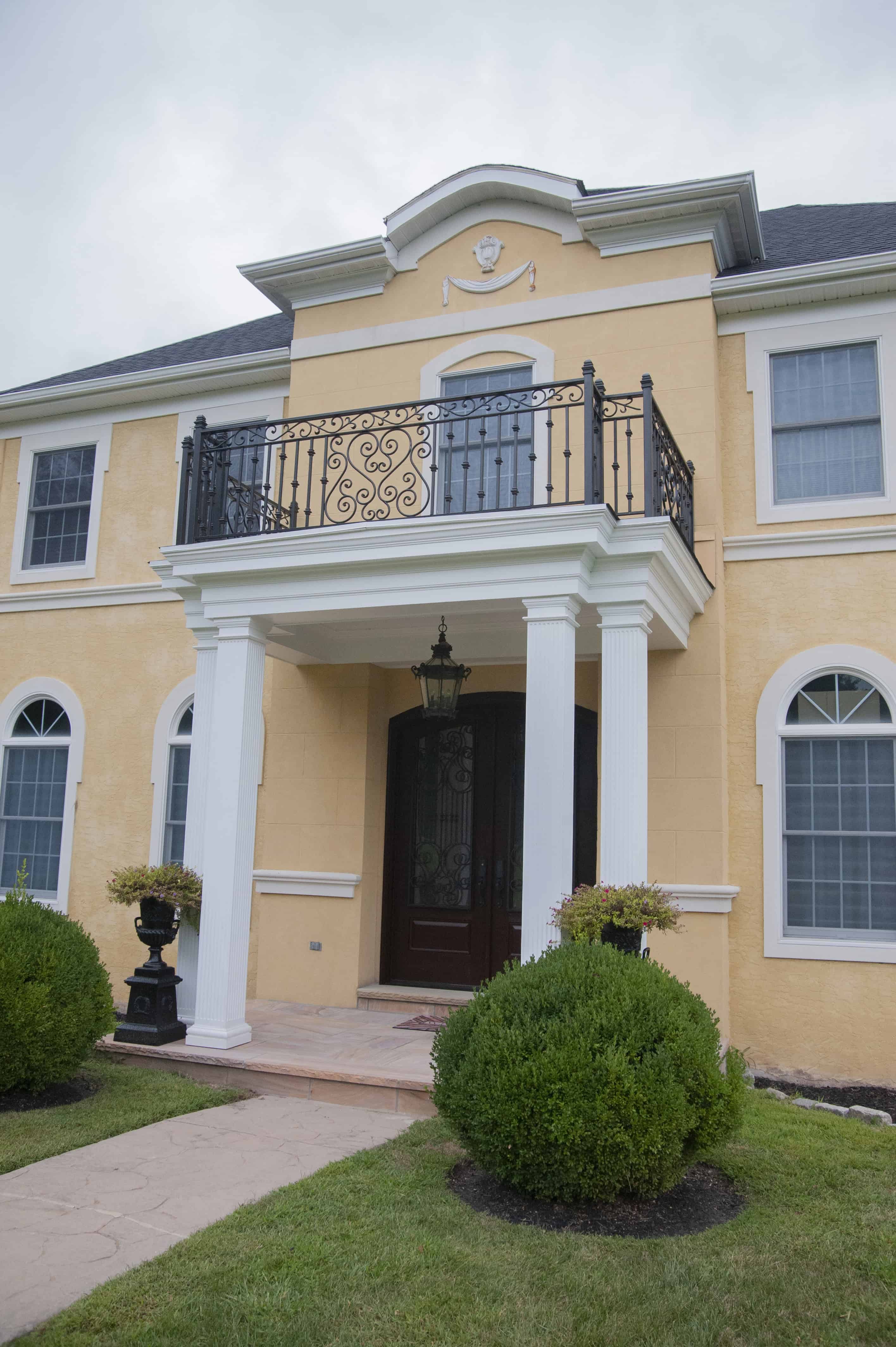 A yellow two-story house with white columns, a black ornate balcony, arched windows, and neatly trimmed shrubs along a stone walkway leading to the front door.