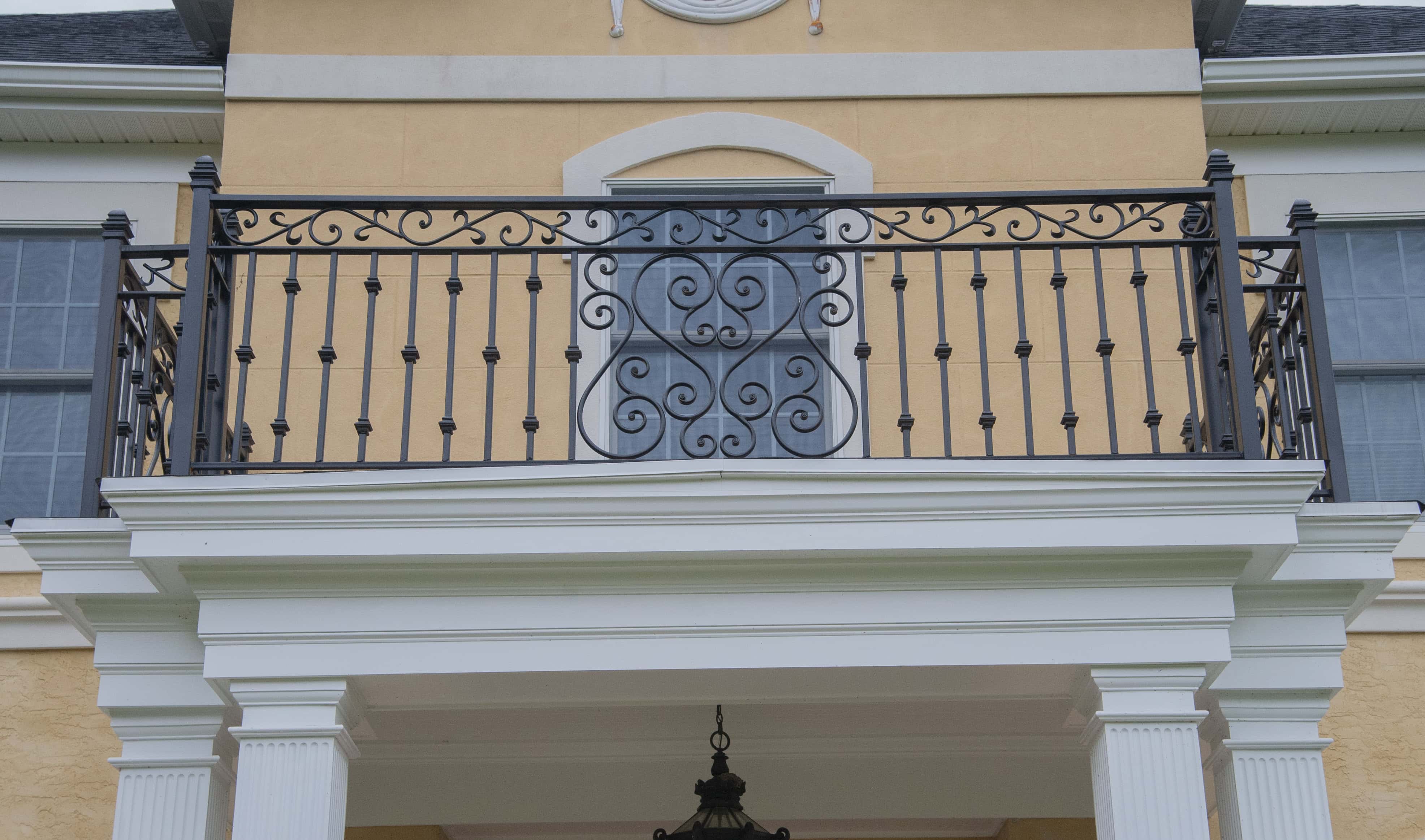 A black wrought iron balcony with ornate scrollwork designs sits above white columns on a yellow building with blue-gray window shutters.