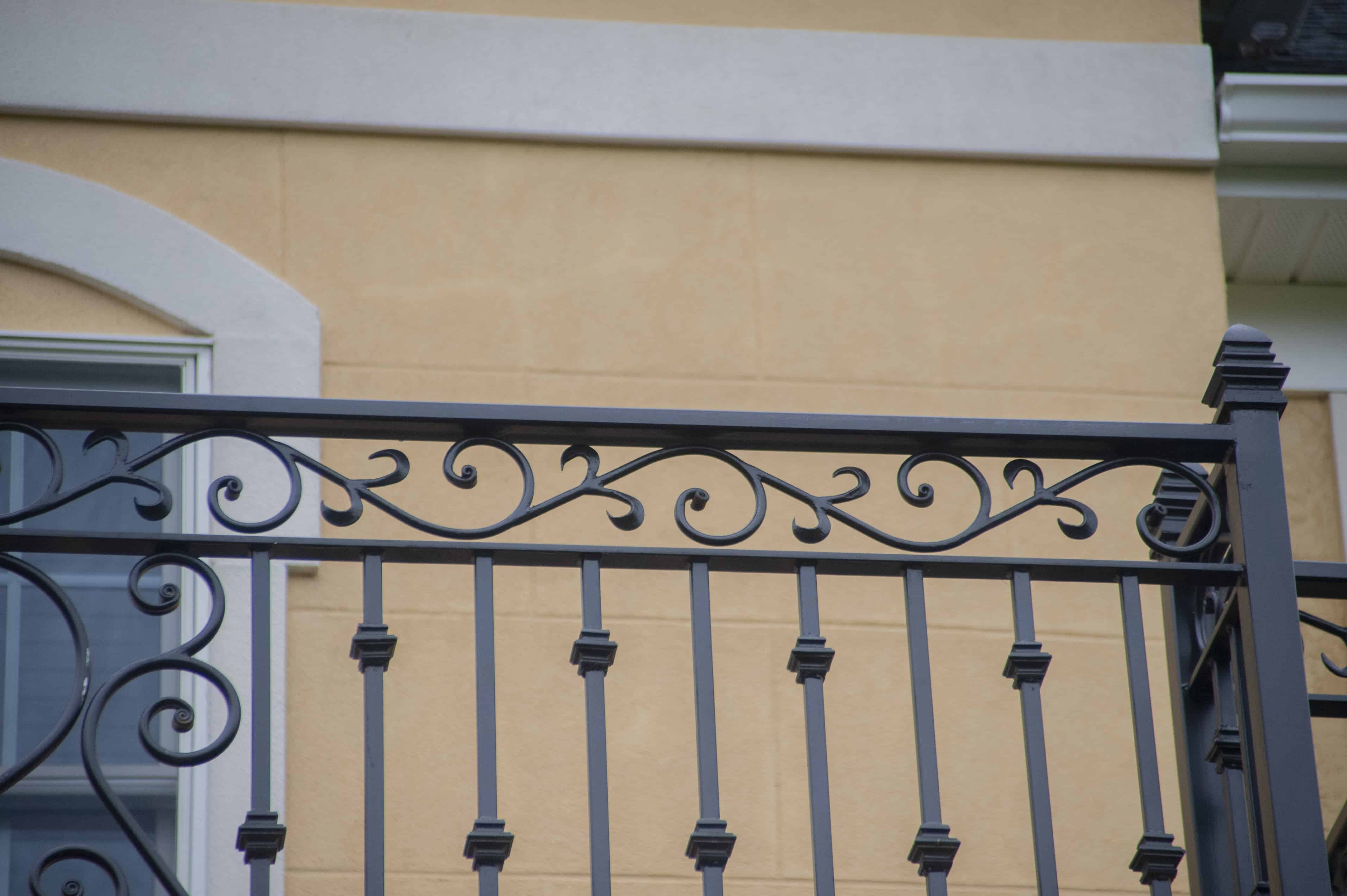 Black wrought iron balcony railing with decorative scrollwork patterns in front of a beige stucco building facade. A window with a white frame is partially visible on the left side.