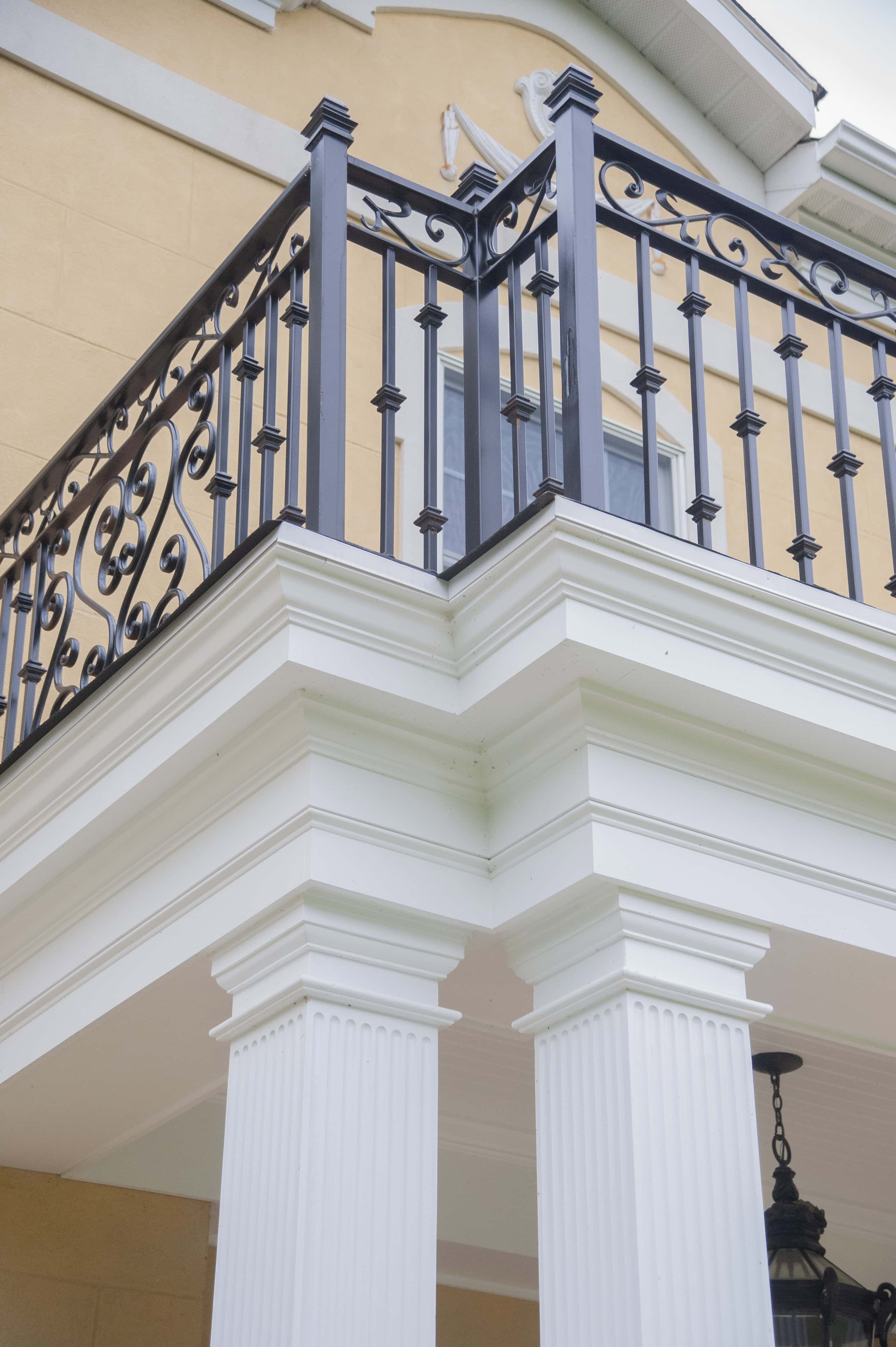 Close-up of a building’s corner with white columns and trim, supporting a balcony with ornate black metal railings; part of a window and beige exterior wall are visible above.