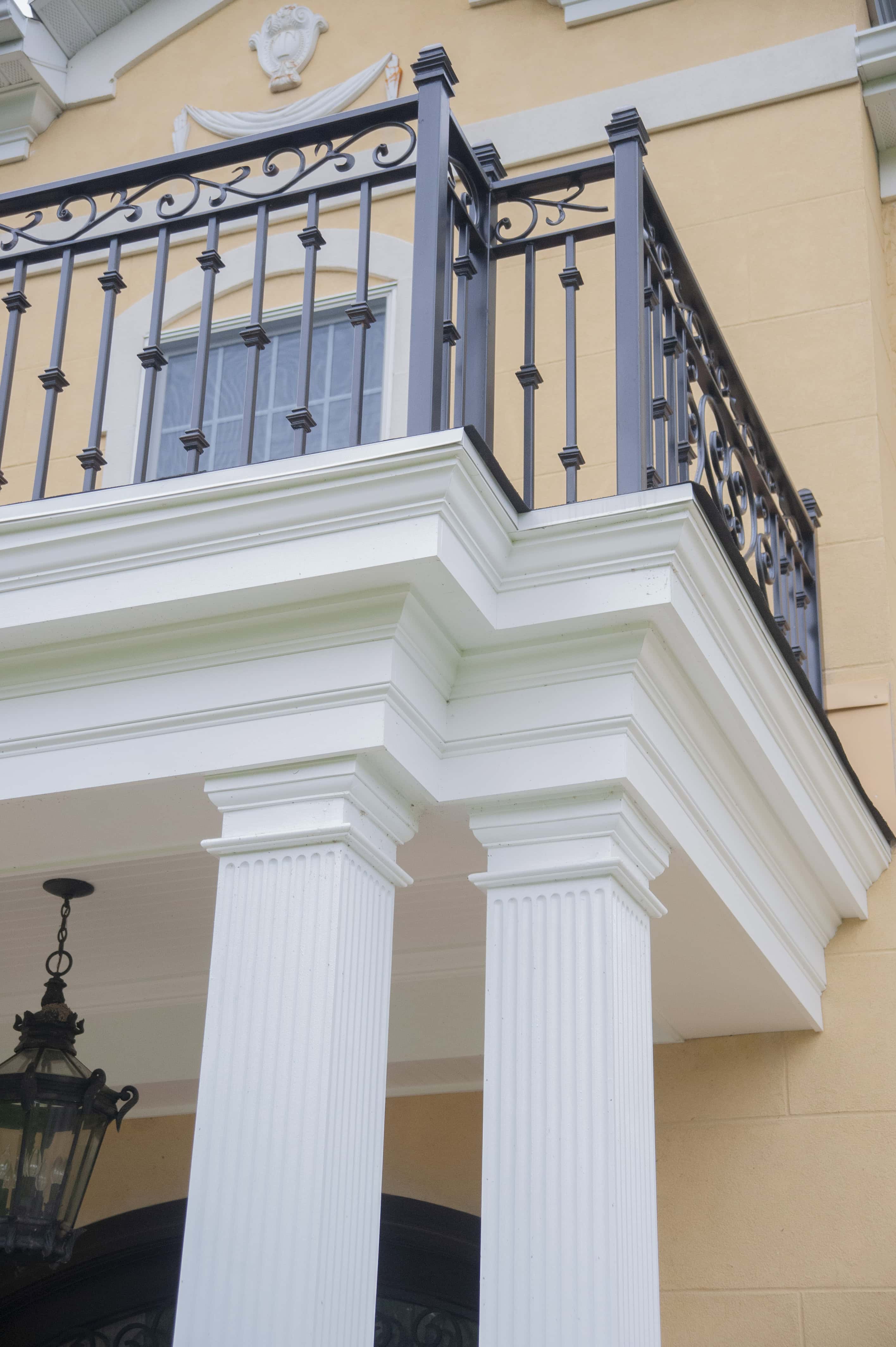 Two white, fluted columns support a balcony with an ornate black metal railing on a tan building. A black lantern hangs from the ceiling above the entrance below.