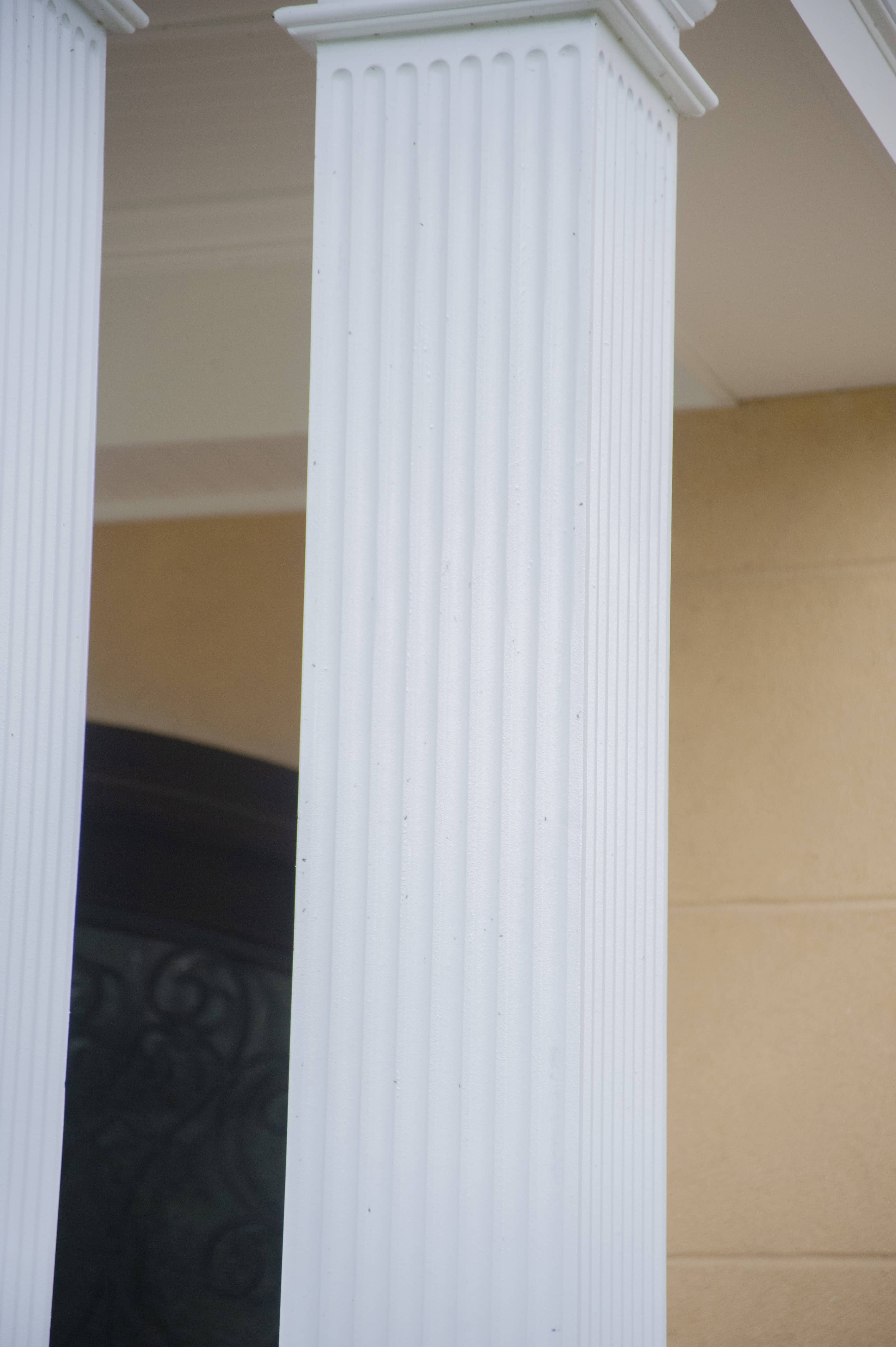 A close-up of a white, fluted column with vertical grooves, supporting a building with a beige wall and part of a dark arched window visible in the background.