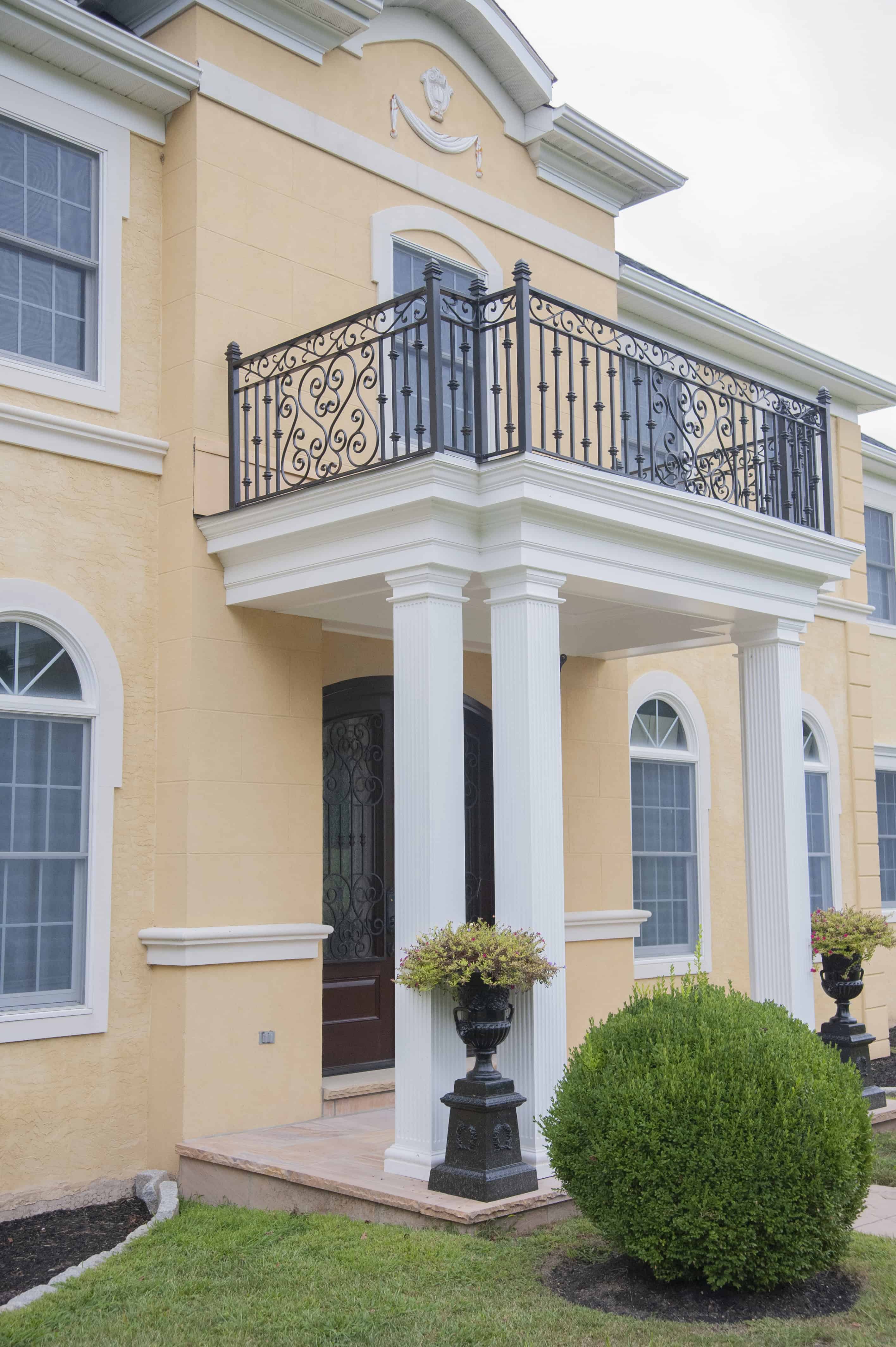 A yellow two-story house with white columns, large arched windows, and a decorative black wrought iron balcony above the front entrance. Two black planters with greenery are on either side of the entry walkway.