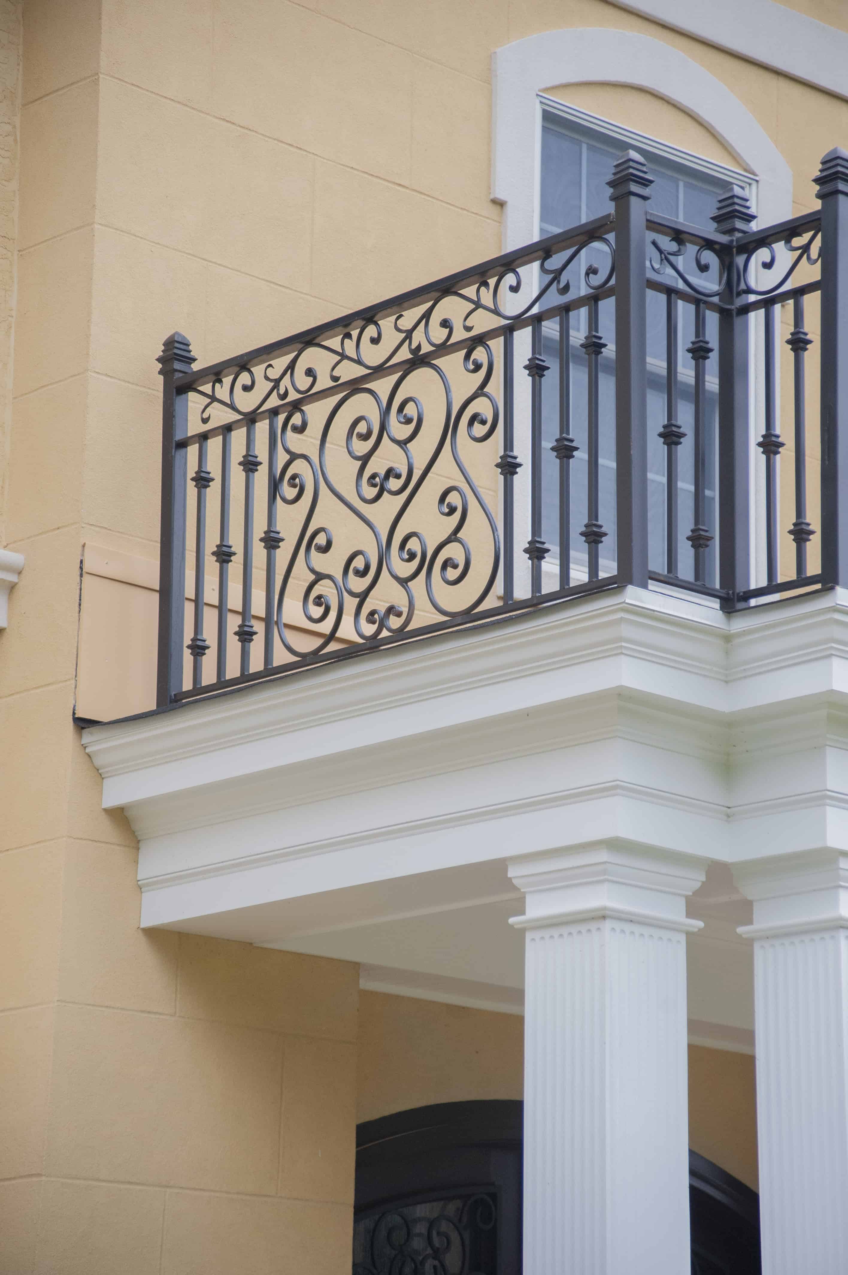 A close-up of a building’s exterior shows a decorative wrought iron balcony railing with intricate scrollwork, above white columns and a yellow stucco wall, next to a window with an arched top.