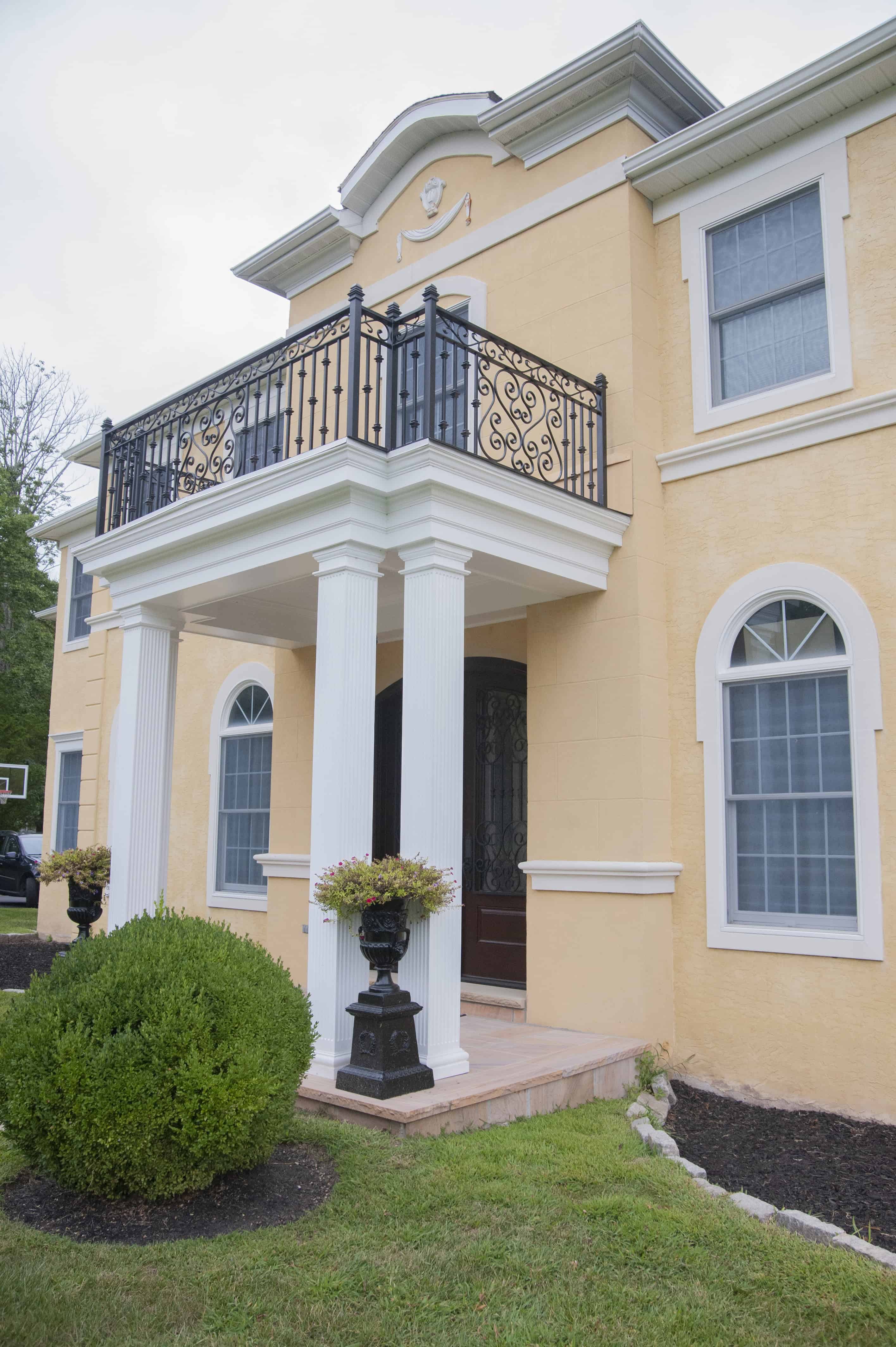 A two-story yellow stucco house with white columns, a decorative black wrought iron balcony, large windows, and landscaped shrubs in the front yard.