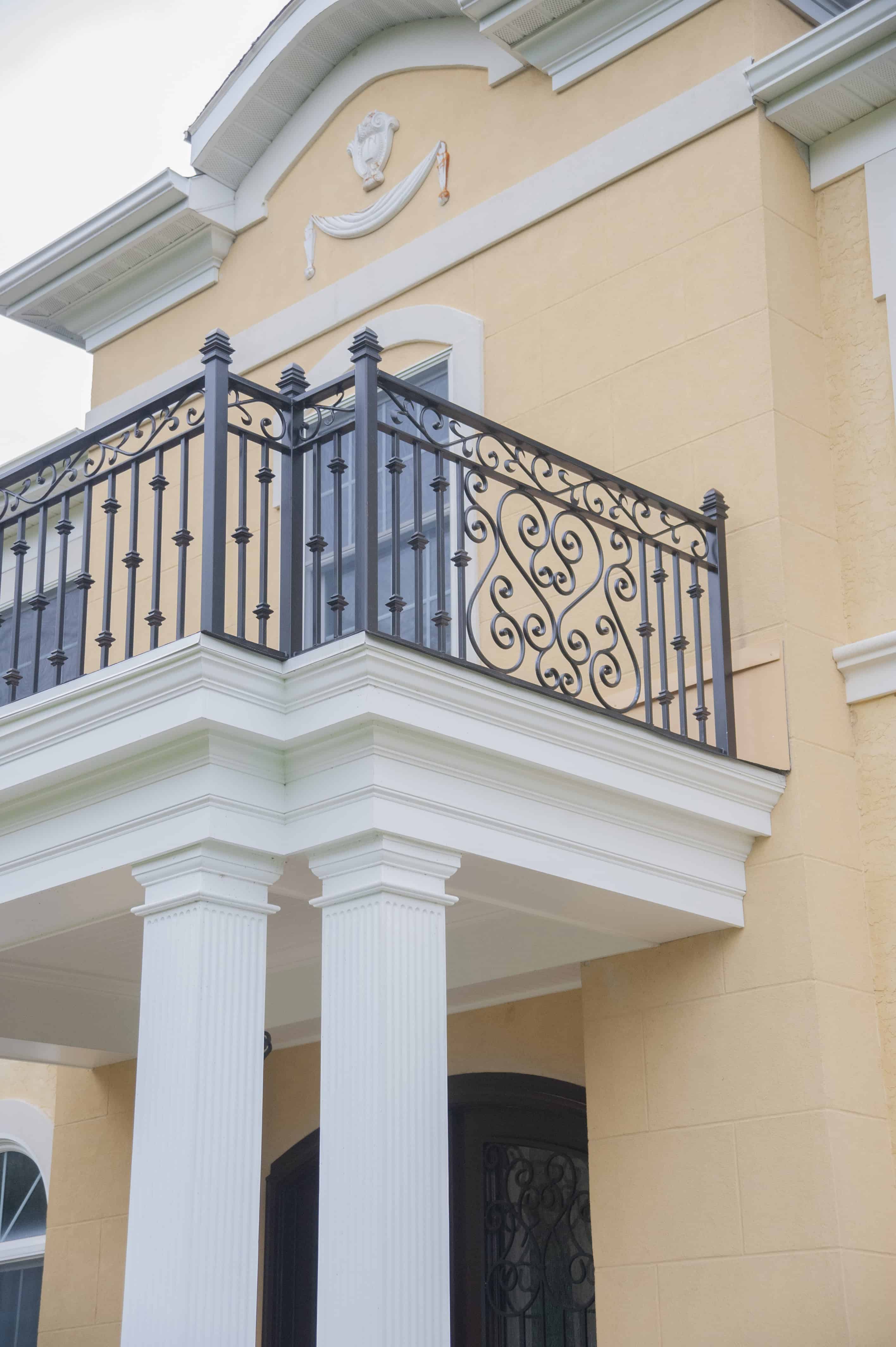 A close-up of a yellow building’s exterior shows white columns supporting a balcony with ornate black wrought iron railings and decorative trim under a light gray roof.