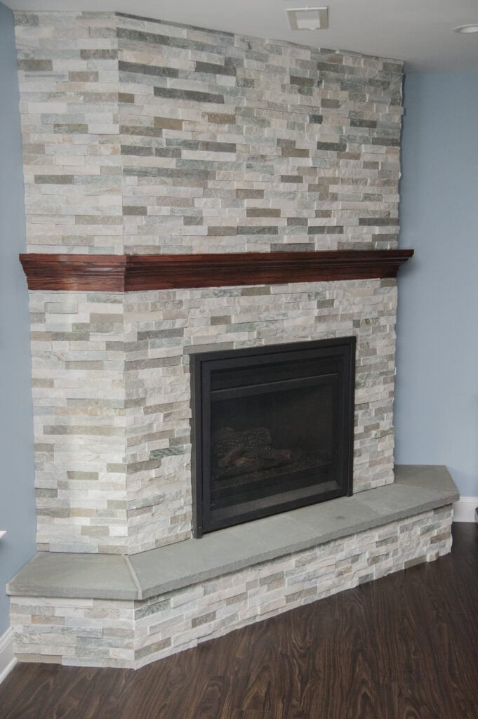 A modern fireplace with a rectangular black insert is set in a wall covered with light gray and white stone tiles. A dark wooden mantel runs across the middle, and the floor is dark wood. The walls are painted light blue.
