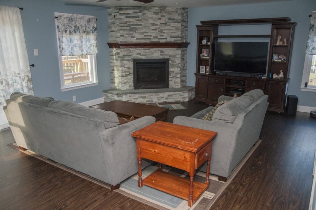 A cozy living room with two gray sofas facing a stone fireplace and a large TV in a dark wood entertainment center, wooden coffee and side tables, and bright windows with patterned curtains.