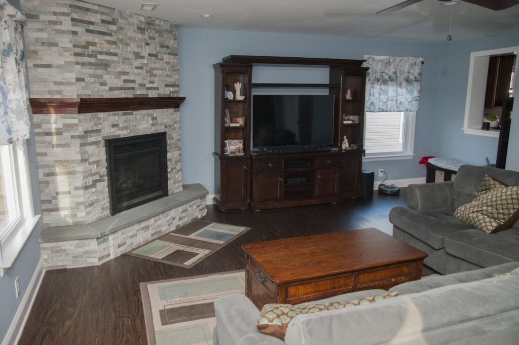 A living room with blue walls, a stone fireplace, a large wooden entertainment center with a TV, a gray sofa with patterned pillows, a wooden coffee table, and two small rugs on a dark wood floor.