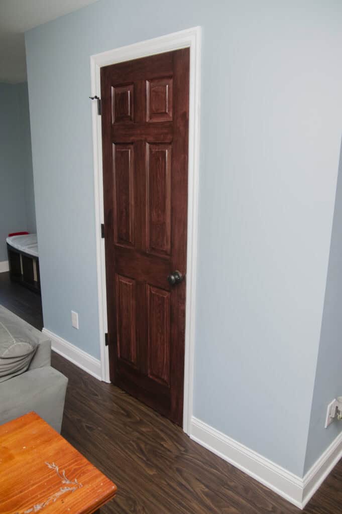 A narrow, dark wooden door with a metal knob set in a light blue wall, next to a gray couch and wood floor, with a corner of an orange table visible in the foreground.