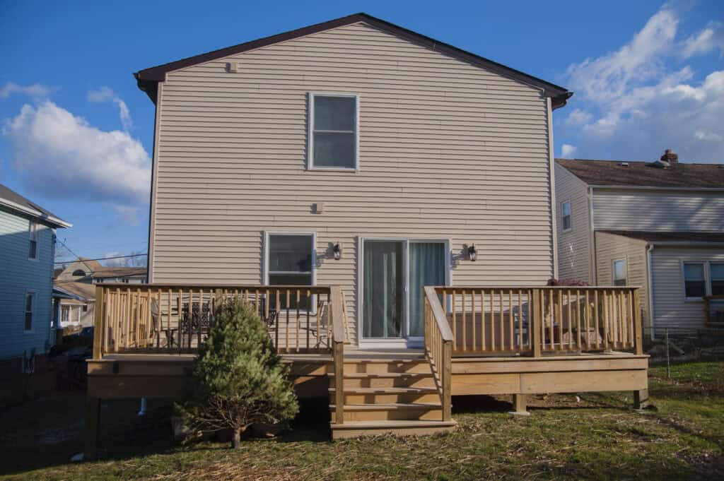 A beige two-story house with a wooden deck, railing, and steps leading to a backyard. A small evergreen shrub sits to the left of the stairs. Blue sky and neighboring houses are visible.