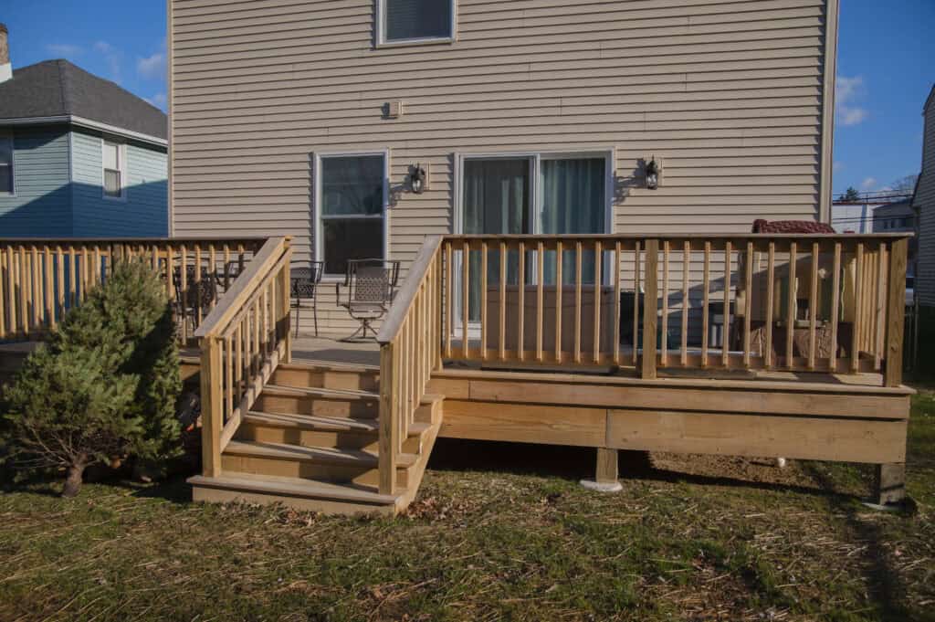 A newly built wooden deck with stairs and railings attached to the back of a beige house, next to a small evergreen shrub and grassy yard.
