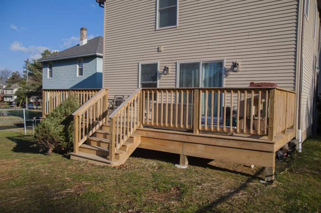 A newly built wooden deck with railings and steps is attached to the back of a beige house, overlooking a grassy backyard. Two windows and outdoor lights are visible on the house.
