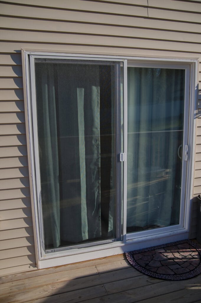 A closed glass sliding door with light blue curtains behind it, set in a tan vinyl-sided wall. Part of a round doormat and wooden deck are visible at the bottom of the image.