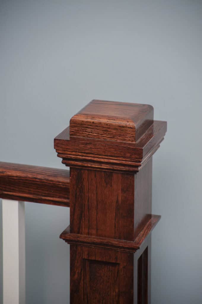 A close-up of a polished wooden staircase newel post with a square cap, set against a light blue wall. White balusters are partially visible to the left.