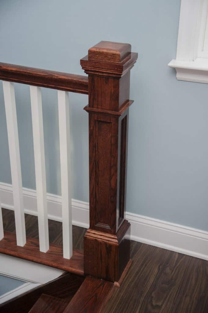 A dark wood staircase newel post with white balusters and a matching handrail, set against a light blue wall and window, with dark wood flooring and white baseboard trim.