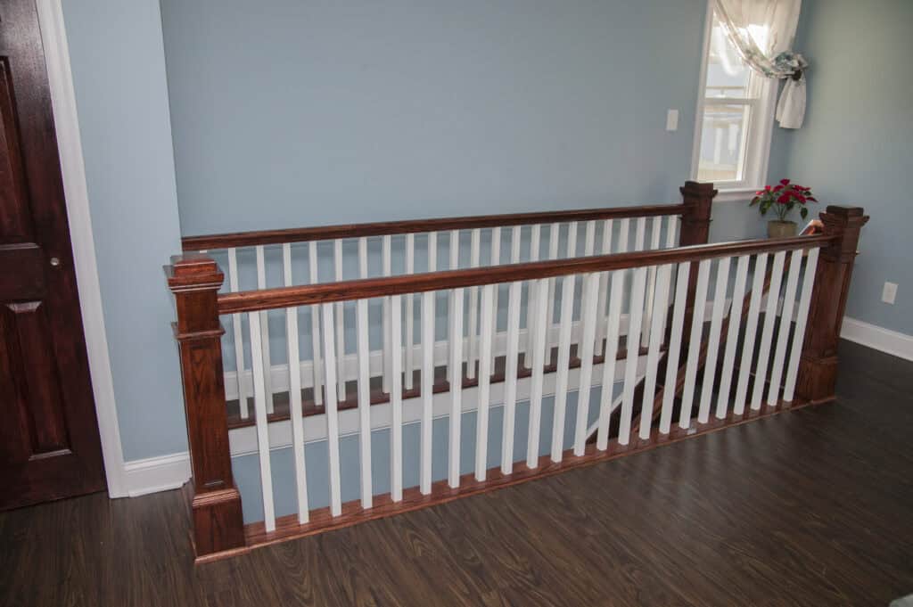A wooden stair railing with white vertical balusters and dark brown handrails stands on a polished dark wood floor in a light blue-walled room with a window and a small table with a plant.