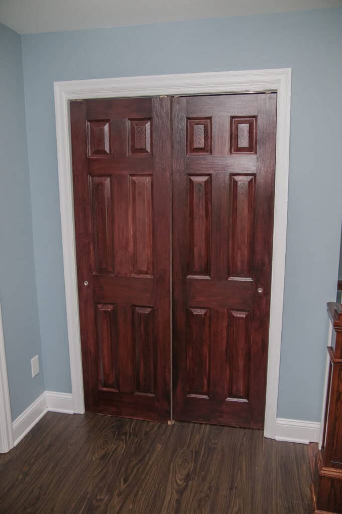 Two dark wood closet doors with white trim are set into a blue wall above dark wood flooring in a room. The doors have a classic panel design and are closed.