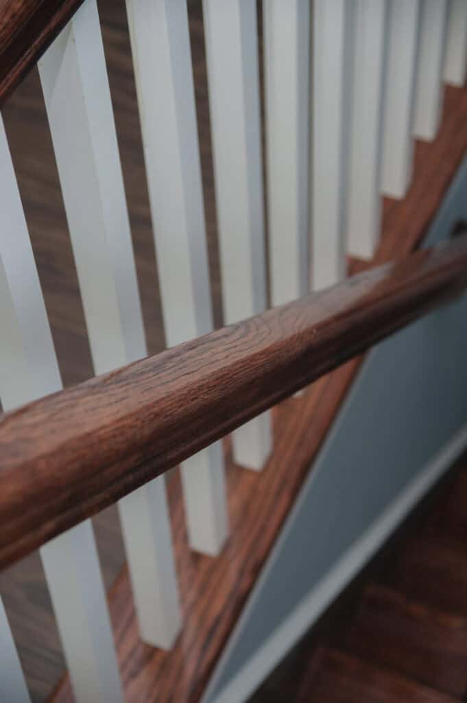 Close-up view of a wooden staircase with a polished dark wood handrail and white balusters, leading downward, with part of the stair treads and blue wall visible.
