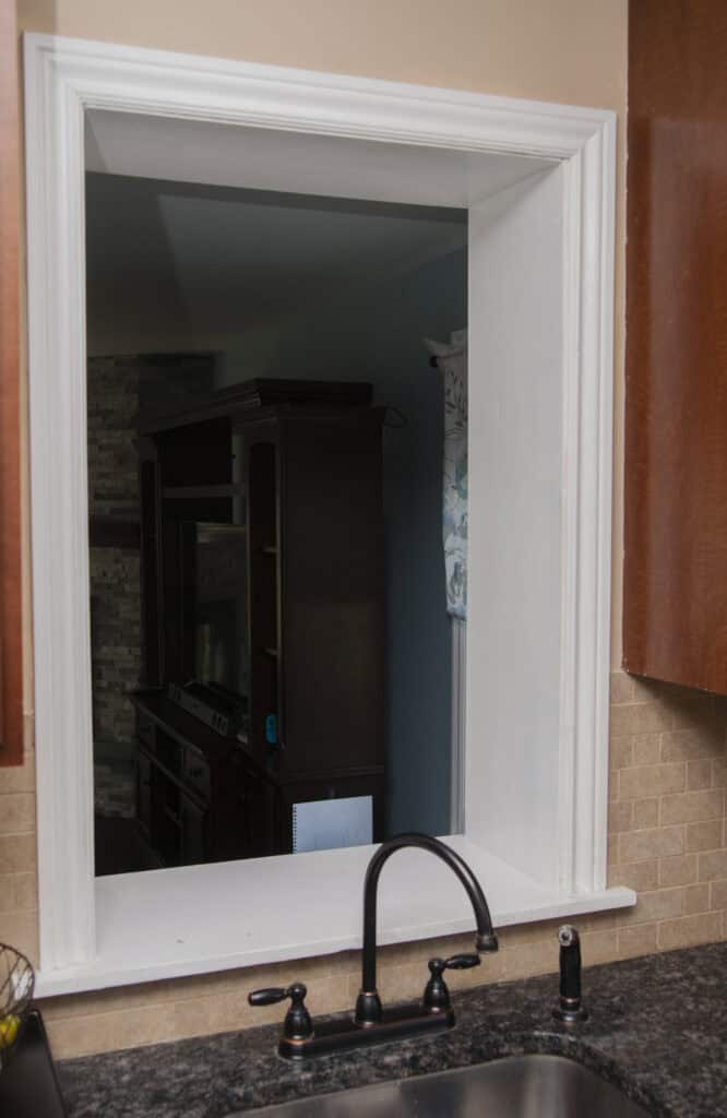 A kitchen sink with a black faucet is set below a large, open passthrough window that looks into a dimly lit living room with a dark entertainment center and stone accent wall.
