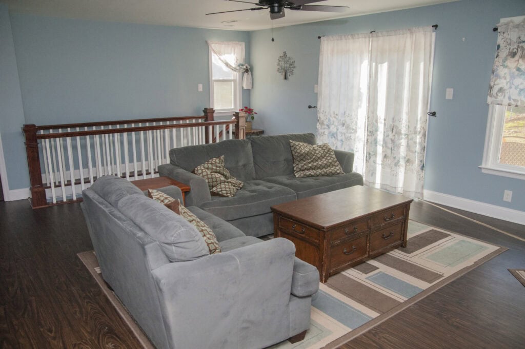 A living room with blue walls, two gray sofas, a wooden coffee table, patterned pillows, an area rug, hardwood floors, and sheer curtains on large windows letting in natural light.