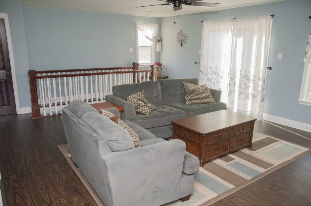 A living room with two gray sofas, a wooden coffee table, patterned pillows, a striped rug, wood flooring, blue walls, white curtains, and a wooden railing by the stairwell.