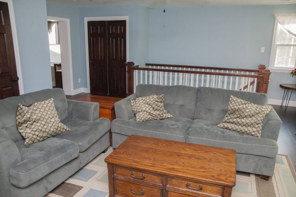 A cozy living room with two gray sofas, patterned throw pillows, a wooden coffee table, hardwood floors, light blue walls, and dark wooden doors. Sunlight streams in through a window with white curtains.