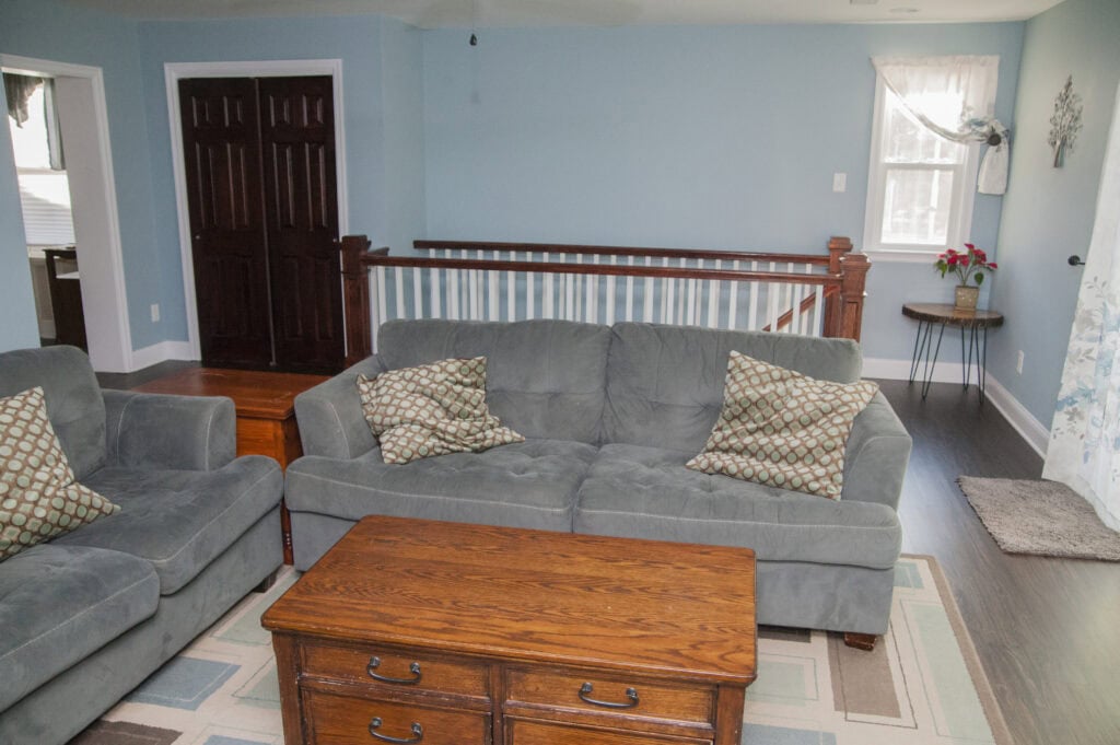 A living room with light blue walls, a gray couch and armchair with patterned pillows, a wooden coffee table, a striped area rug, and a side table with a vase of red flowers near a window.