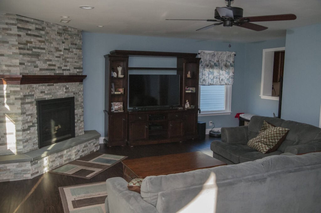 A living room with a stone fireplace, large TV on a dark wood entertainment center, gray couches, a wooden coffee table, blue walls, and windows with floral curtains. Sunlight shines on the wooden floor.