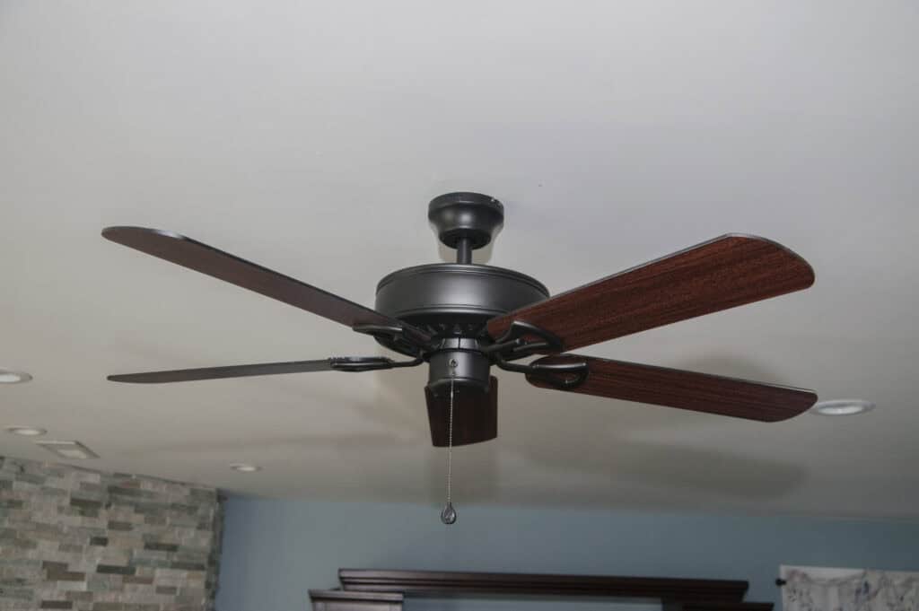 A ceiling fan with four dark wooden blades and a black center is mounted on a white ceiling in a modern room with stone and wood accents.