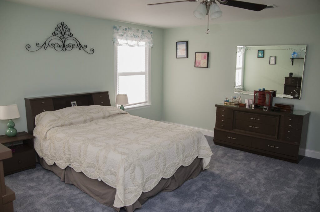 A tidy bedroom with a bed covered in a patterned white quilt, brown wooden furniture, a dresser with a mirror, a window with a white valance, wall art, and blue carpeted flooring.