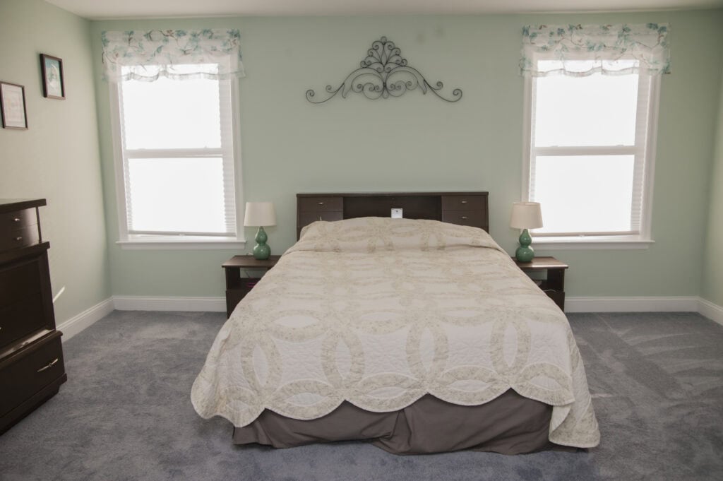 A neatly made bed with a patterned beige quilt is centered between two windows in a light green bedroom. Each side has a nightstand with a green lamp. The floor is carpeted, and there is minimal decor.