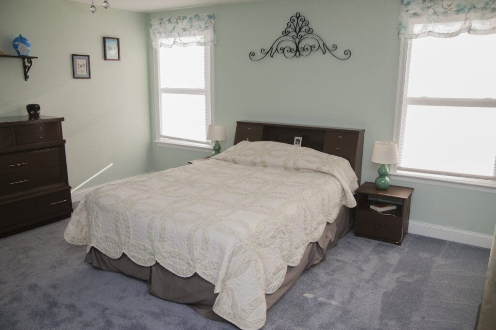 A neatly made bed with a cream quilt and two green lamps on bedside tables sits between two windows in a light-filled bedroom with pale walls, blue carpet, and dark wood furniture.
