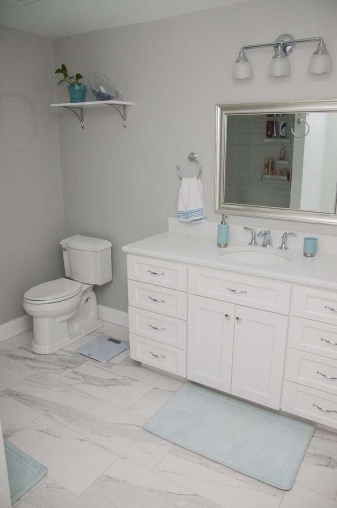 A modern bathroom with white cabinets, a sink, a large mirror, and silver fixtures. There is a toilet, two light blue rugs, a shelf with plants and decor, and light gray walls with marble-patterned tile flooring.