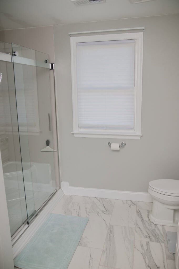 A clean, modern bathroom with a glass-enclosed shower, white toilet, marble tile floor, light gray walls, a window with a white blind, and a light blue bath mat.