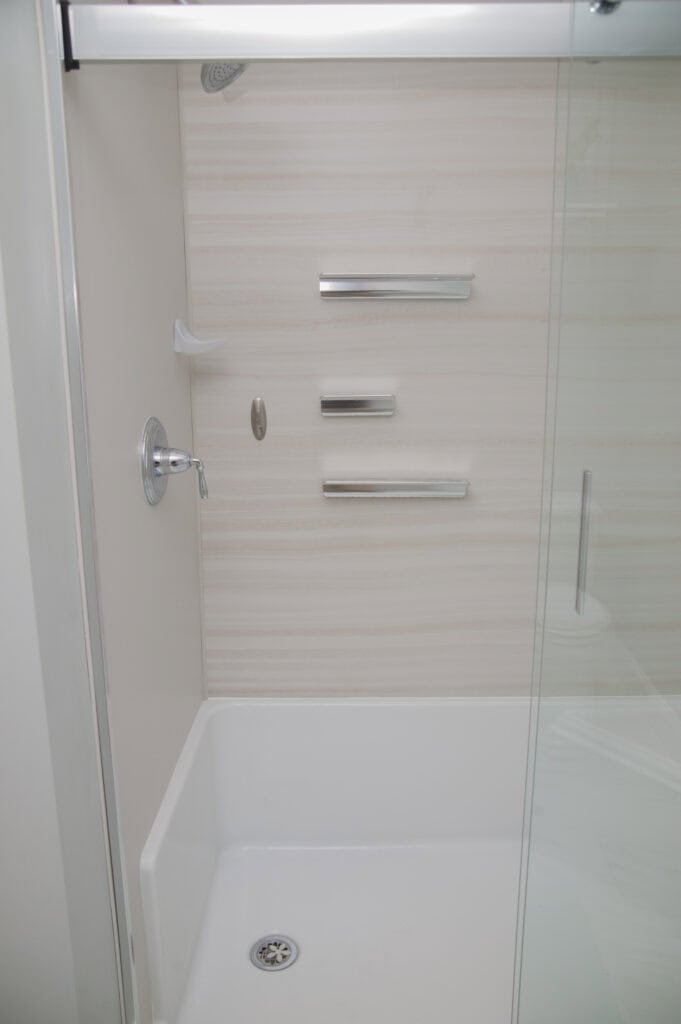 A modern shower with a glass door, silver shower handle, three built-in metal shelves, a white acrylic base, and light-colored wall panels.