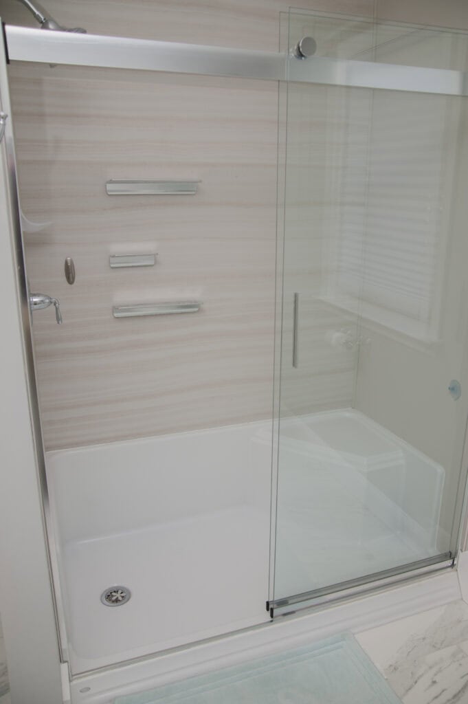 A modern shower with light-colored walls, three metal shelves, and a sliding glass door. The shower has a white base, a silver drain, and a blue bath mat outside on the floor.