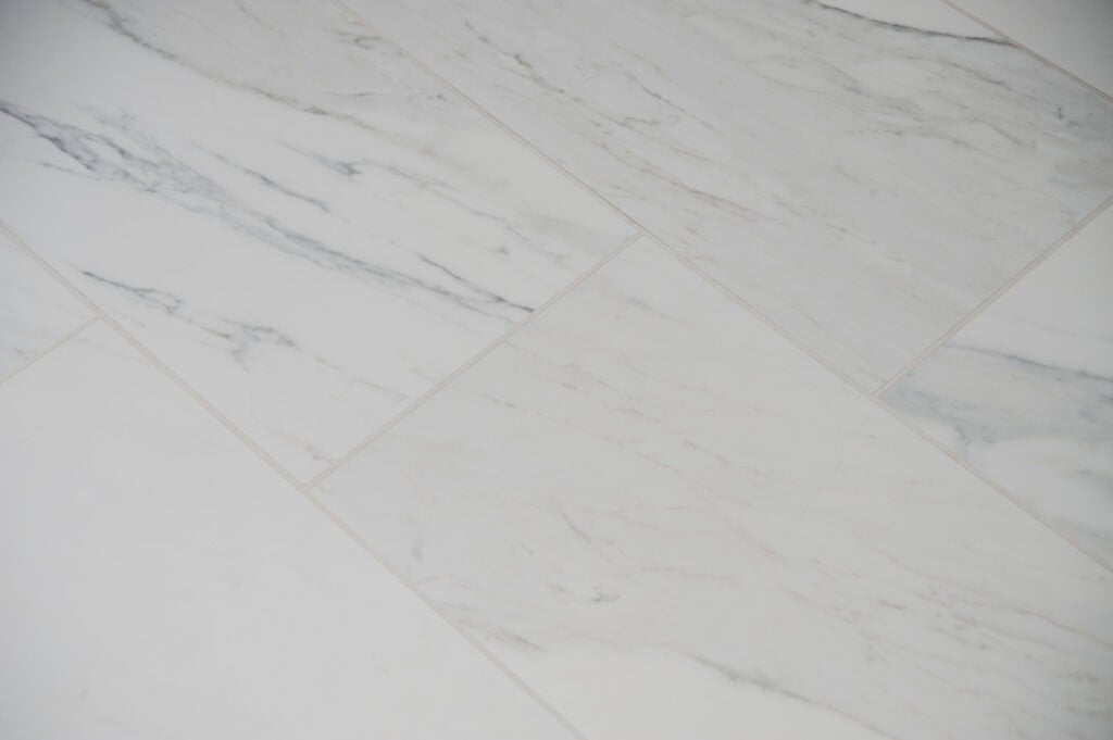 Close-up view of white marble floor tiles with subtle gray veining, arranged in a staggered pattern. The surface appears smooth and polished, giving a clean and elegant look.