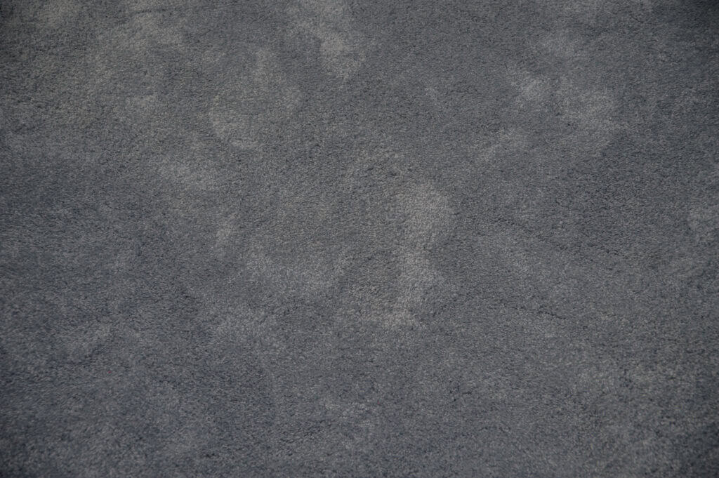 Close-up view of a grey carpet with subtle texture and irregular patterns, showing variations in shading and pile direction.