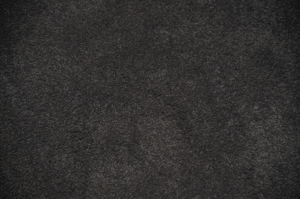 A close-up view of a dark grey, textured surface, possibly a fabric or carpet, with a slightly uneven and mottled appearance.