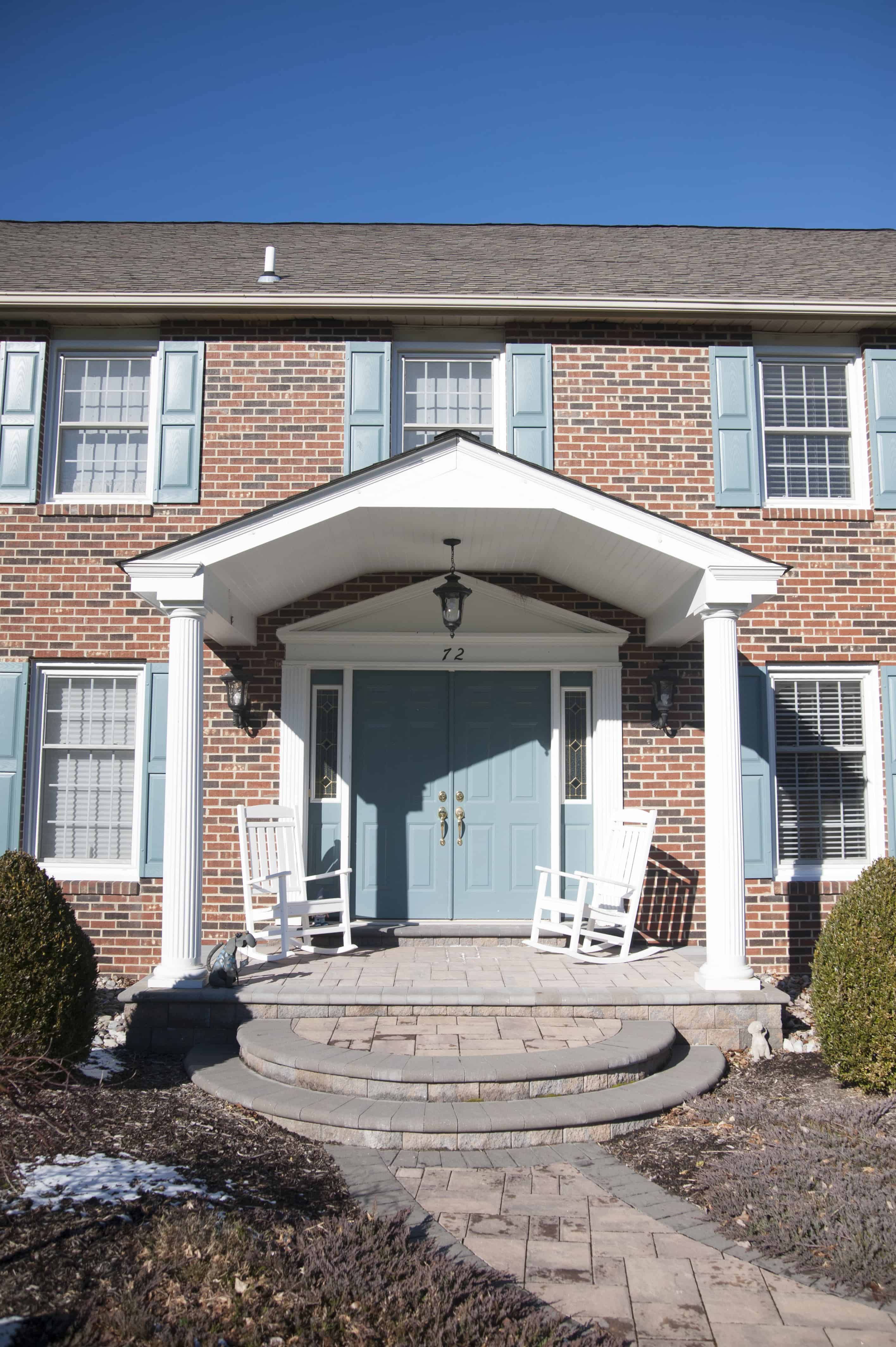 A red brick house with blue shutters and a covered front porch, featuring two white rocking chairs by the double-door entrance and a clear blue sky overhead.