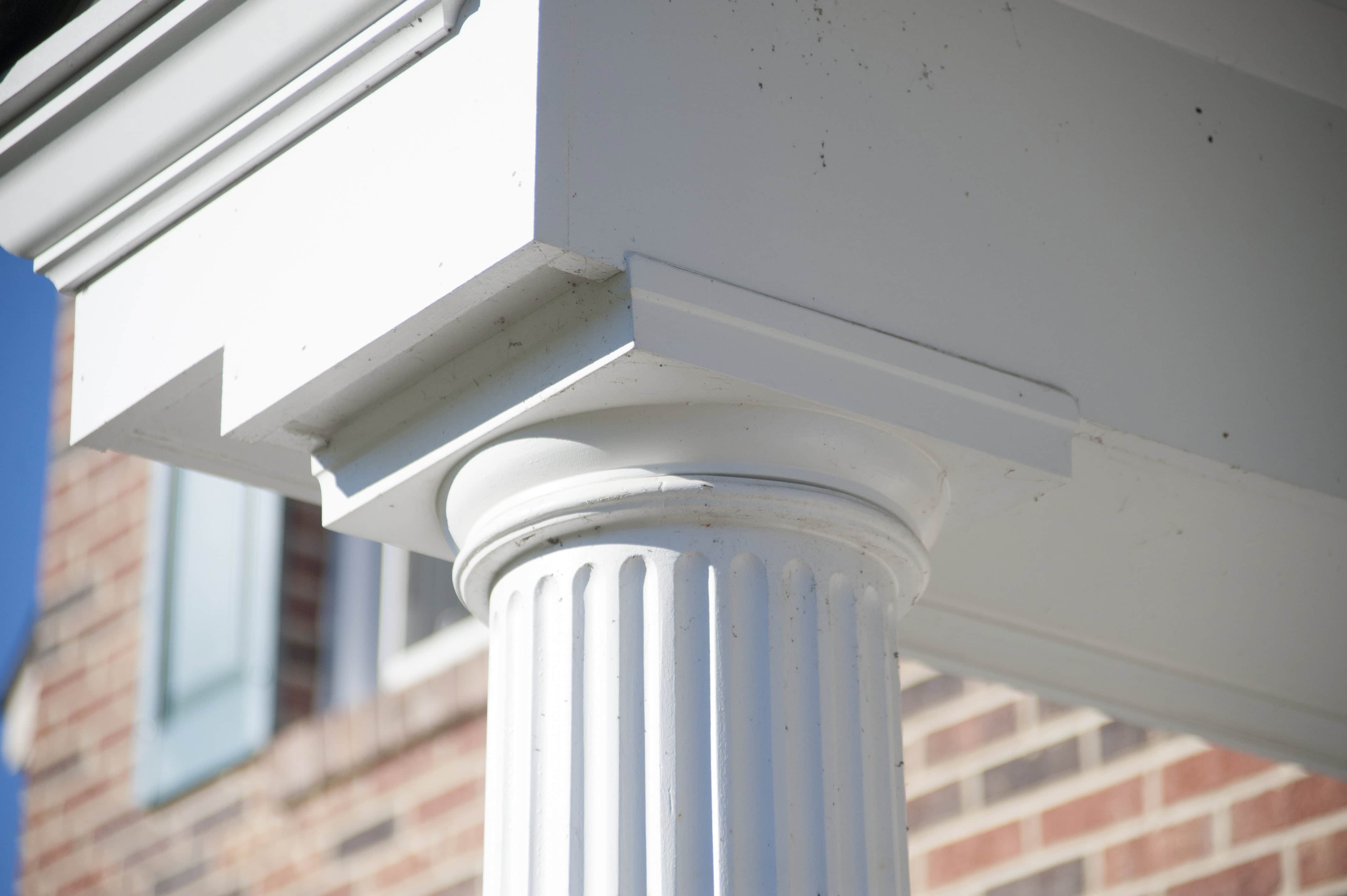 Close-up of a white fluted column supporting a porch roof, with a brick building and blue window shutter visible in the blurred background.