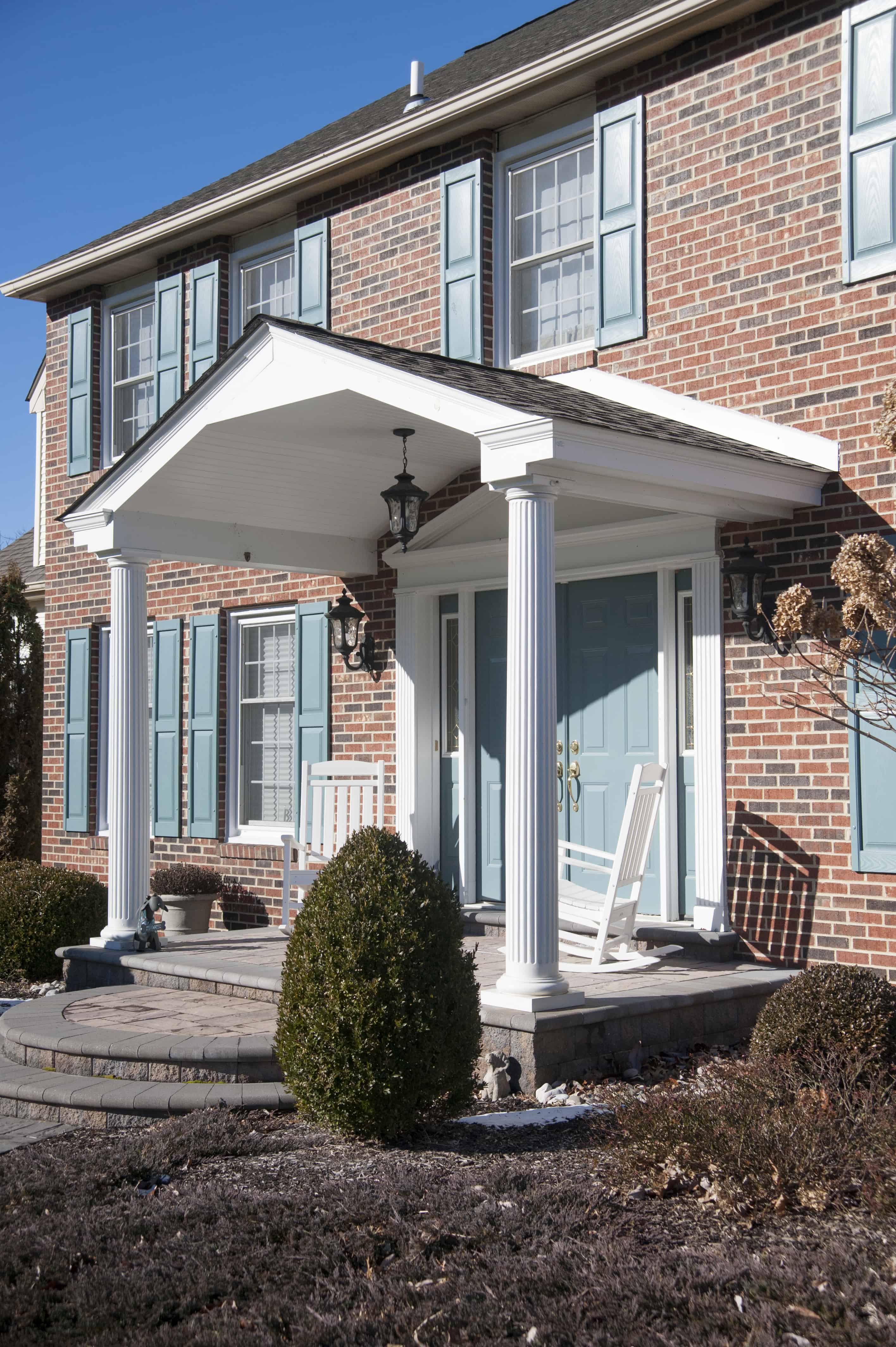 A red brick house with blue shutters and double front doors features a white columned porch with rocking chairs and a walkway, surrounded by trimmed bushes under a clear blue sky.