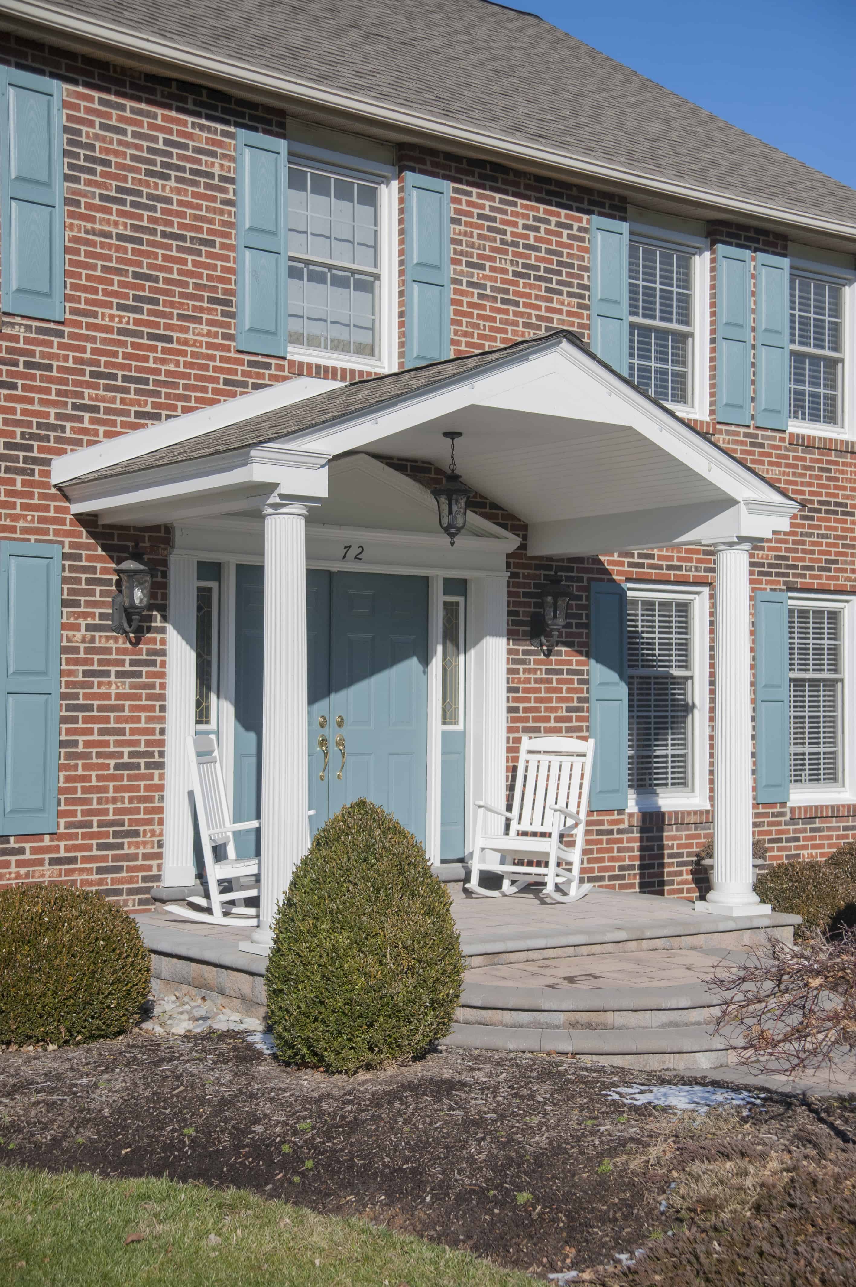 A brick house with blue shutters, white pillars, and a covered front porch. Two white rocking chairs sit on the porch near double doors. Sunlight casts shadows on the steps and trimmed bushes line the walkway.
