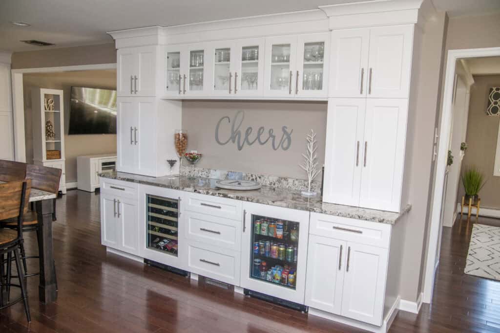 A modern home bar with white cabinets, glass doors, granite countertop, and built-in coolers filled with drinks. Decorative jars and glasses are displayed, with “Cheers” written on the wall above.