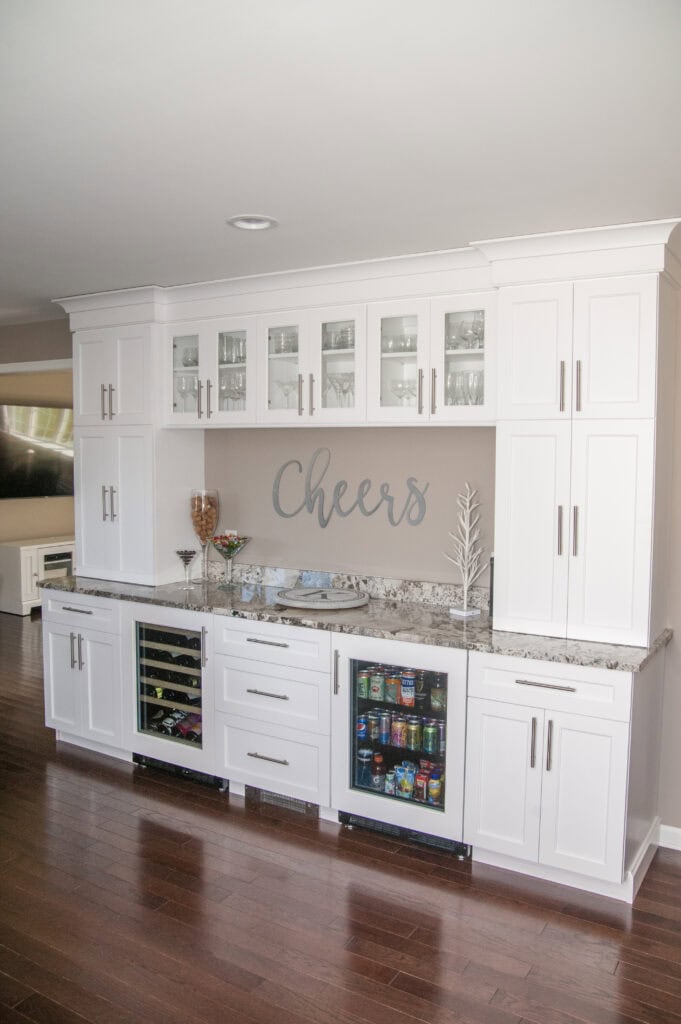 A modern home bar with white cabinets, glass doors, a granite countertop, wine and beverage fridges, snacks, a white decorative tree, and the word Cheers on the wall.